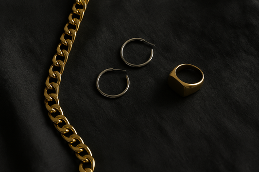 Flat lay of solid 18k gold jewellery including a chunky gold chain, minimalist white gold hoops, and a gold signet ring on a dark textured linen surface, photographed in soft natural light.