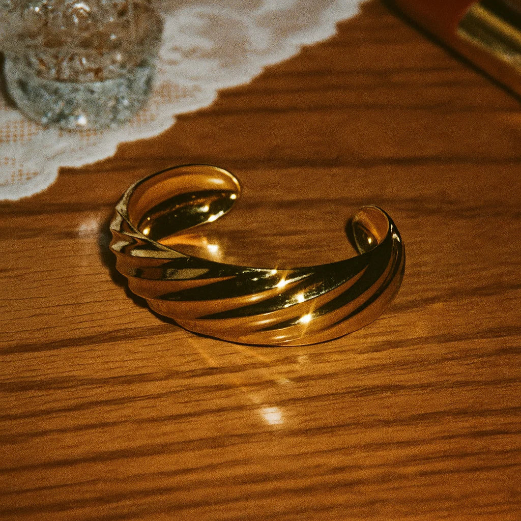 Gold bracelet on a wooden surface with a glass and doily in the background
